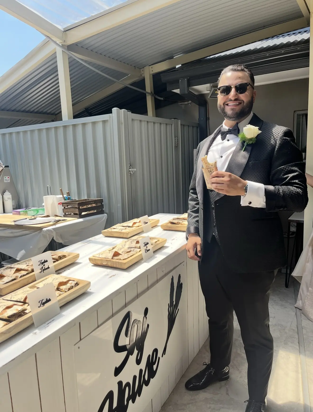 Man in a tuxedo holding a drink at an outdoor food table.