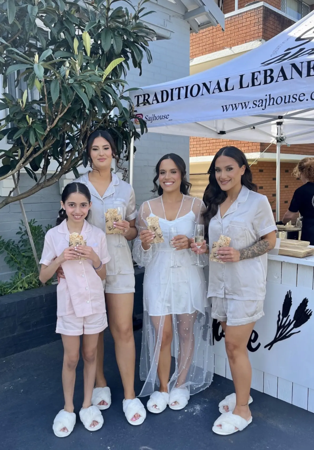 Four women in white and pink outfits holding glasses at an outdoor event.