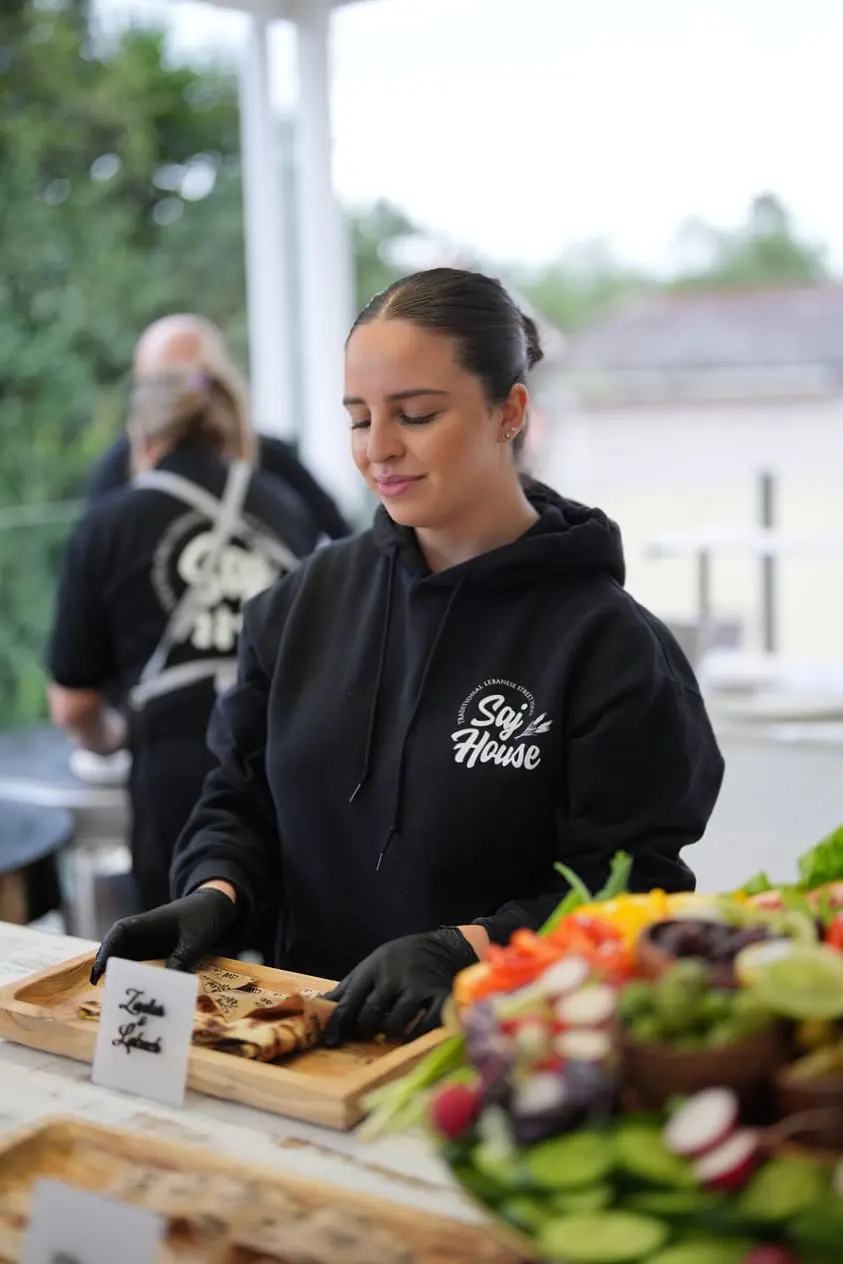 Woman preparing food at an outdoor market stall, wearing a black hoodie.