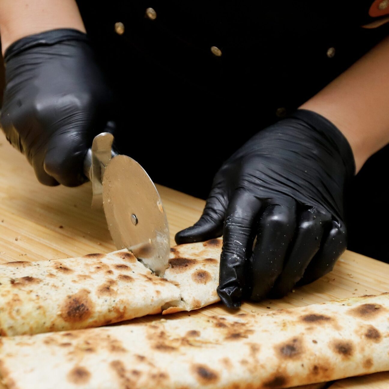 Person slicing a rolled flatbread with a pizza cutter while wearing black gloves.