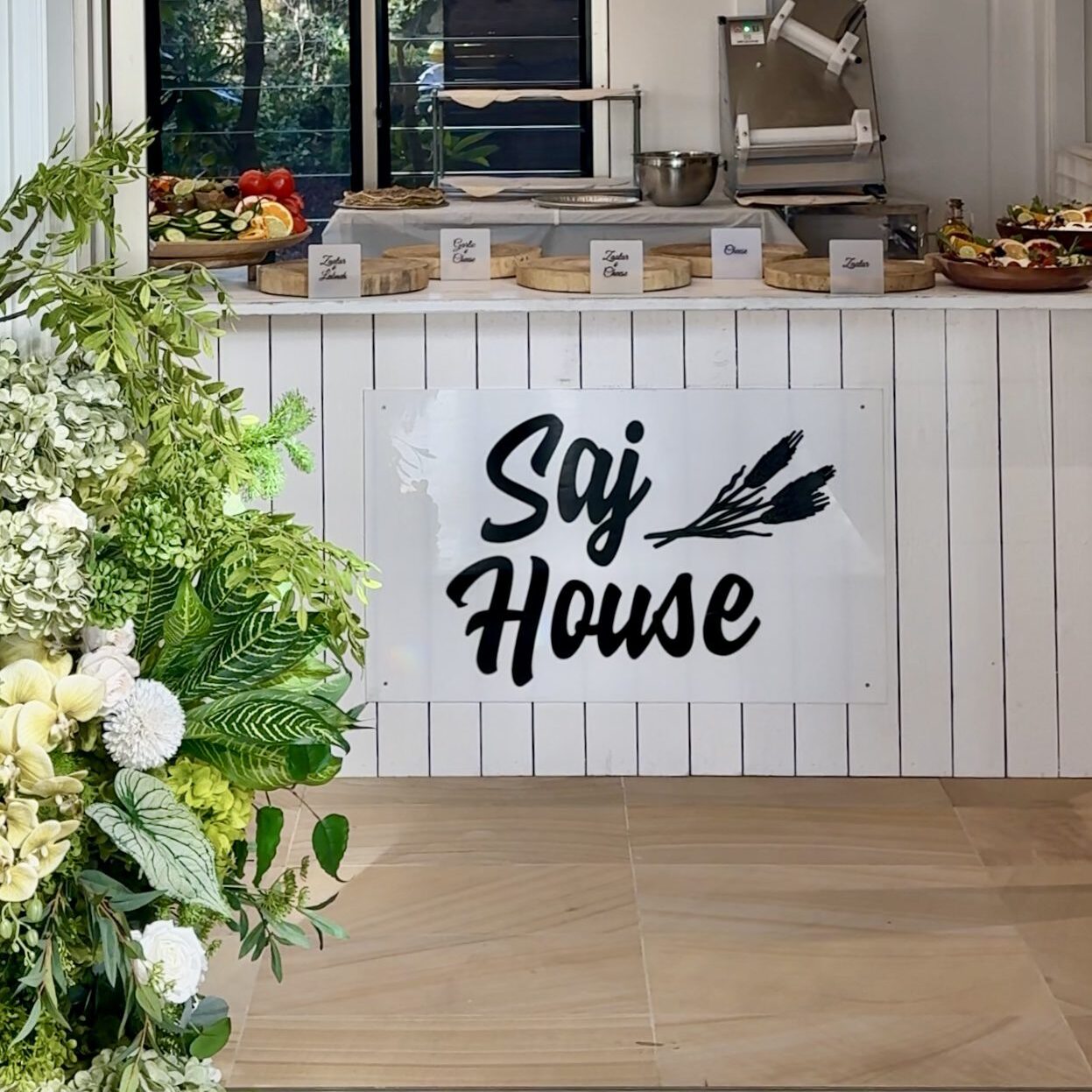 Cozy sign reading 'Sgj House' on white paneled wall with flowers nearby.